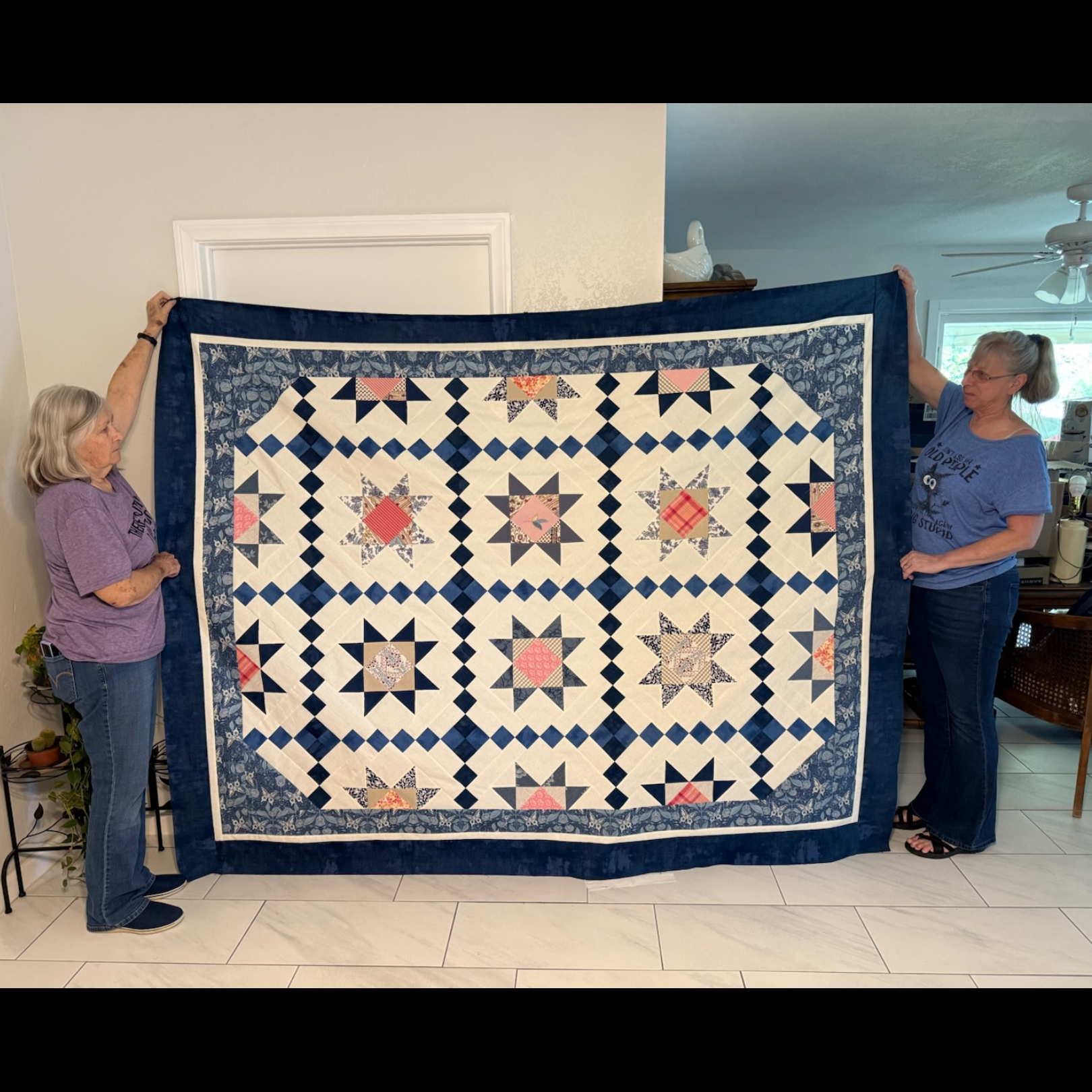Two women proudly display a handmade quilt with floral patterns.